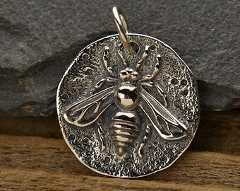 Peut inclure: Breloque en argent avec un motif d'abeille détaillé. L'abeille est centrée sur un disque rond avec une surface texturée et une bordure décorative de petits cercles.