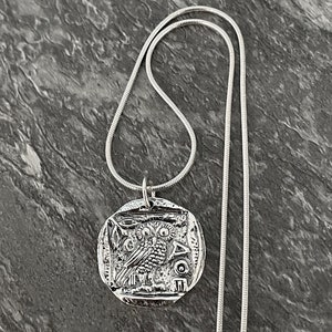 May include: A silver necklace with a round pendant featuring an owl design. The pendant is detailed with intricate carvings and the Greek letters "AOE".