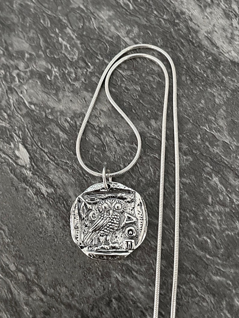 May include: A silver necklace with a round pendant featuring an owl design. The pendant is detailed with intricate carvings and the Greek letters "AOE".