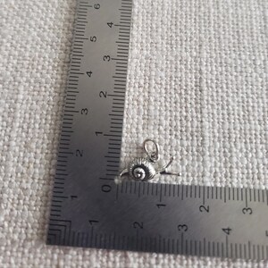 May include: A silver snail charm with a detailed shell and a curled tail. The charm is about 1 inch long and is measured against a ruler.