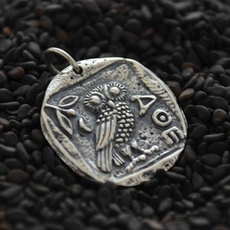 May include: Silver pendant featuring an owl with detailed etching and the Greek word "&Alpha;&Theta;&Eta;&Nu;&Alpha;" (Athena) inscribed around it.