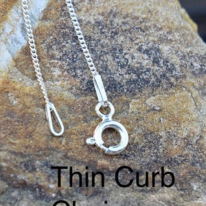 May include: A silver thin curb chain necklace with a lobster clasp and a jump ring. The chain is lying on a textured rock surface.