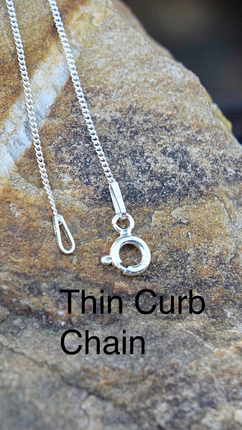 May include: A silver thin curb chain necklace with a lobster clasp and a jump ring. The chain is lying on a textured rock surface.