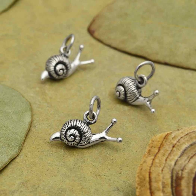 May include: Three silver snail charms with detailed shell and body features. The charms are on a light brown background.