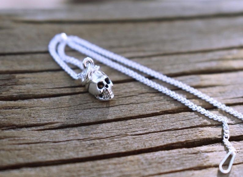 May include: A silver chain necklace with a small silver skull pendant. The skull has a detailed design and is attached to the chain with a small loop.