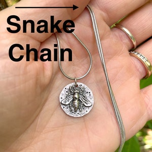 May include: A silver snake chain necklace with a round pendant featuring a detailed bee design.