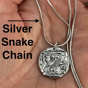 May include: Silver snake chain necklace with a round pendant featuring a detailed engraving of an owl. The pendant has the inscription "AOE" on the bottom.