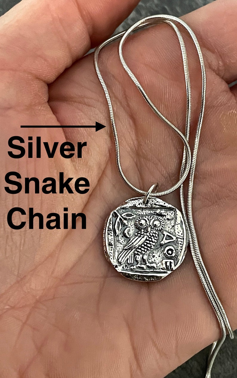 May include: Silver snake chain necklace with a round pendant featuring a detailed engraving of an owl. The pendant has the inscription "AOE" on the bottom.