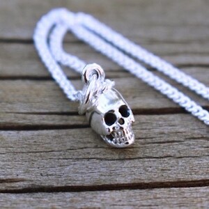 May include: A silver chain necklace with a small silver skull pendant. The skull is detailed with eye sockets and a jawline.