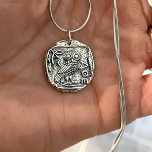 May include: Silver necklace with an owl pendant. The pendant is a round coin with an owl and the word "AOE" on it.