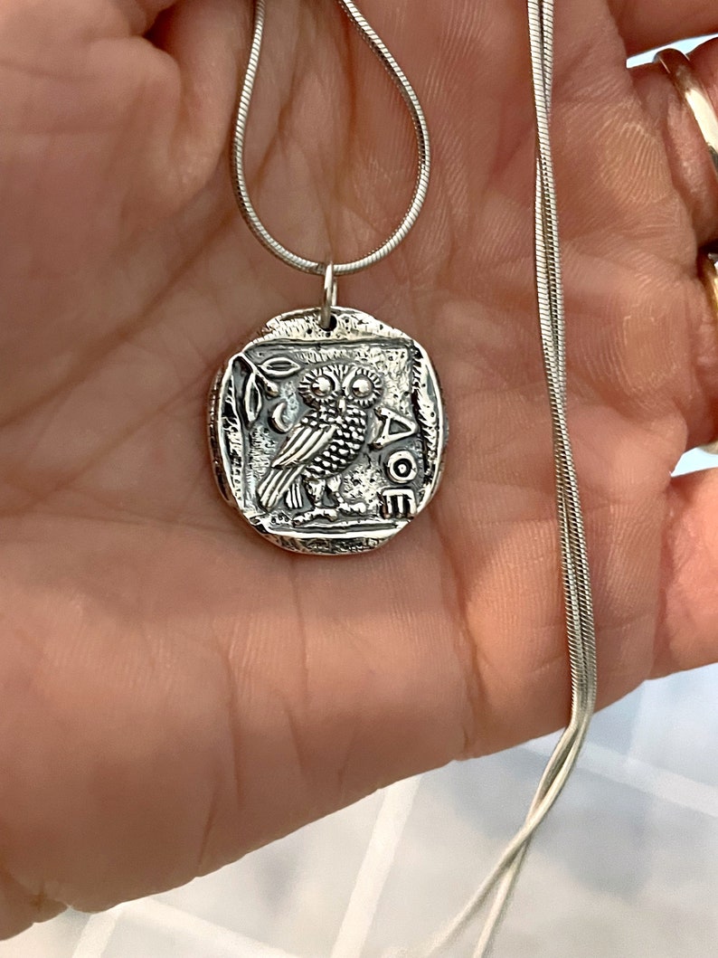 May include: Silver necklace with an owl pendant. The pendant is a round coin with an owl and the word "AOE" on it.