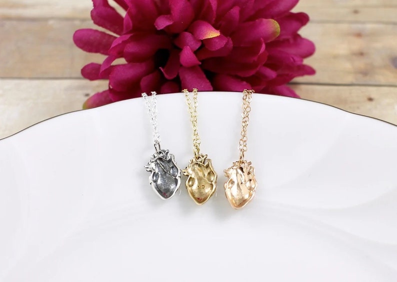 May include: Three silver, gold, and rose gold necklaces with anatomical heart pendants. The necklaces are displayed on a white surface.