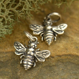 May include: Two silver bee charms with detailed wings and bodies. The charms are on a brown textured surface.