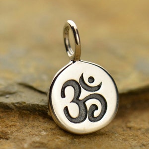 May include: A silver-colored pendant with the Om symbol. The pendant is circular with a raised border and a loop for a chain. The Om symbol is in a dark color, contrasting with the silver background. The pendant is likely made of metal.