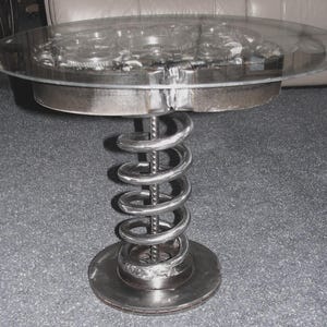 May include: A round glass table top with a metal base made of gears and a large metal spring. The table is silver in color and has a modern industrial design.