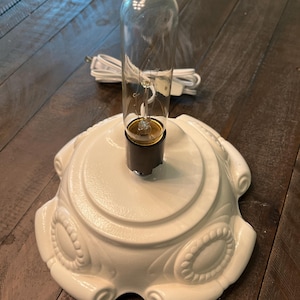 May include: A white, decorative lamp base with a clear glass light bulb. The base has an ornate, raised design and a dark gray socket. A white power cord is visible.