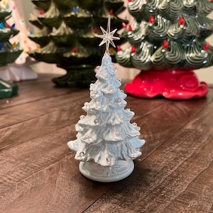 May include: A light blue ceramic Christmas tree with silver glitter accents and clear gem embellishments. The tree is topped with a star and sits on a round base. Other ceramic Christmas trees are in the background.