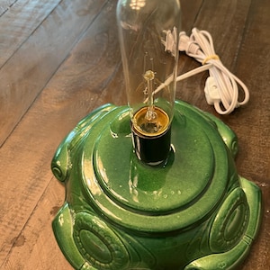 May include: A green ceramic Christmas tree base with a clear lightbulb. The base has a glossy finish and decorative details. A white electrical cord is attached to the base.