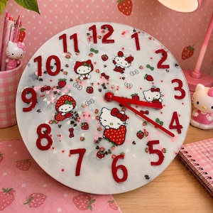May include: A round wall clock with a white marbled background featuring red numbers from 1 to 12. The clock face is decorated with multiple Hello Kitty character illustrations, small red strawberry motifs, and tiny heart-shaped confetti pieces.