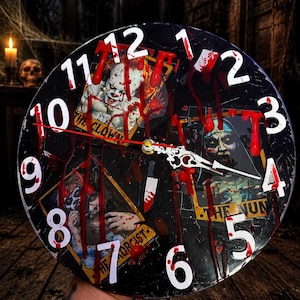 May include: A round wall clock featuring a black background with red blood-like splatters and white numbers. The clock face displays three rectangular horror movie-themed cards labeled The Clown, The Nun, and The Exorcist, with ornate white hands.