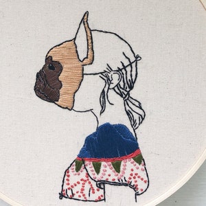 May include: Embroidered hoop art featuring a profile of a person with a dog's head, wearing a blue and green patterned shirt with red polka dots.