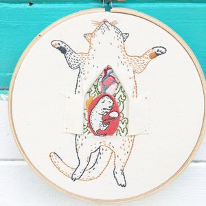 May include: Embroidered hoop art featuring a stylized cat with a smaller cat inside its belly. The larger cat is orange and white with black outlines. The smaller cat is white with black outlines and is curled up inside a red heart. The background is a cream-colored fabric.