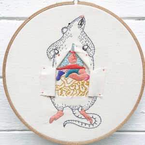 May include: Embroidered hoop art featuring a white rat with its insides exposed, showing colourful organs and a yellow stomach. The rat is standing on its hind legs with its front paws raised.