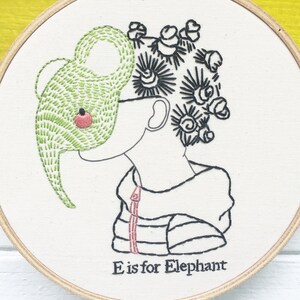 May include: Embroidered hoop art featuring a child with an elephant trunk as a mask. The child has a black and white striped shirt with a red stripe down the front. The text "E is for Elephant" is at the bottom of the hoop.