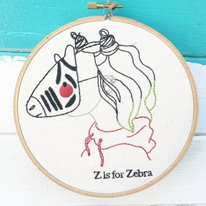 Könnte beinhalten: Gesticktes Stickrahmen-Kunstwerk mit einem Zebra-Design. Das Zebra hat schwarze Streifen, ein rotes Auge und eine rote und grüne Mähne. Der Text "Z is for Zebra" ist darunter gestickt. Der Rahmen ist aus Holz.
