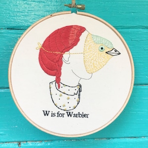以下が含まれることがあります： 赤い髪の女の子が黄色と緑の鳥のマスクをしている刺繍のフープアート。画像の下に「W is for Warbler」というテキストが書かれています。