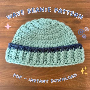 May include: A light mint green crochet beanie with a dark navy blue ribbed brim. The text WAVE BEANIE PATTERN and PDF - INSTANT DOWNLOAD are displayed in white with a blue outline around the beanie on a wooden background.