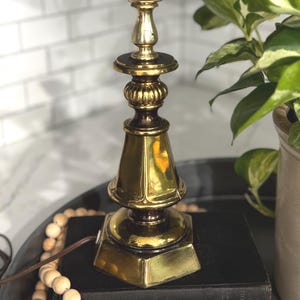 May include: A vintage brass table lamp with a detailed, ornate design. The lamp features a square base, a conical body, and a bulb socket at the top. The lamp is a warm gold color and sits on a dark surface.