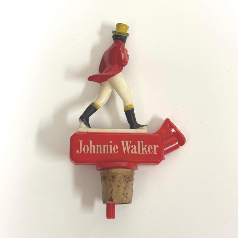 Puede incluir: Un tap&oacute;n de botella rojo con el logotipo de Johnnie Walker y una figura en miniatura del hombre que camina. La figura viste un abrigo rojo, pantalones blancos, botas negras y un sombrero de copa amarillo. El tap&oacute;n tiene una base de corcho.