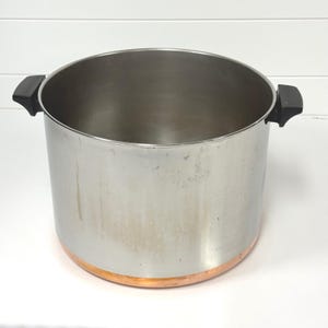 May include: A large stainless steel stockpot with a polished silver finish and a visible copper base. The pot features two black heat-resistant side handles attached to the upper rim, providing a sturdy grip for lifting this heavy-duty kitchen vessel.