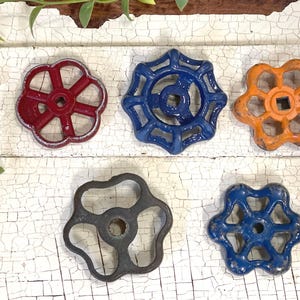 May include: Five vintage water valve handles in various colors: red, blue, orange, gray, and dark blue. Each handle has a unique design with a central square opening and a flower-like shape. The handles are made of metal and show signs of wear.
