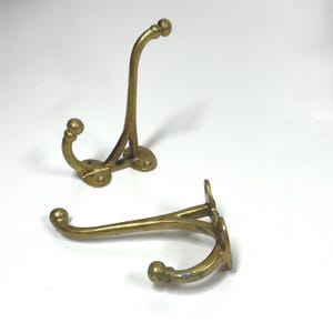 May include: Two brass coat hooks with a polished finish, each featuring a dual-prong design with rounded ball tips at the ends of the hooks. The hooks have a Y-shaped structure with a flat mounting base containing pre-drilled screw holes.