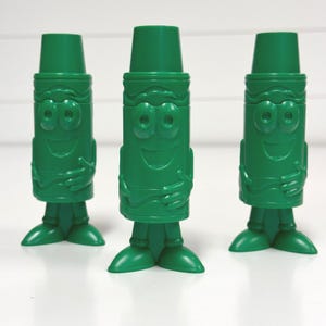 May include: Three green plastic crayon-shaped figures standing upright. Each figure features a molded face with large eyes and a smiling mouth, along with arms resting on the torso and two feet at the base. The design mimics a classic crayon.