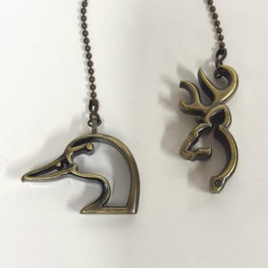 May include: Two antique brass-toned metal ceiling fan pulls attached to beaded chains. One pull is shaped like the profile of a duck head, and the other pull is shaped like the profile of a deer head with antlers, both featuring open-work designs.
