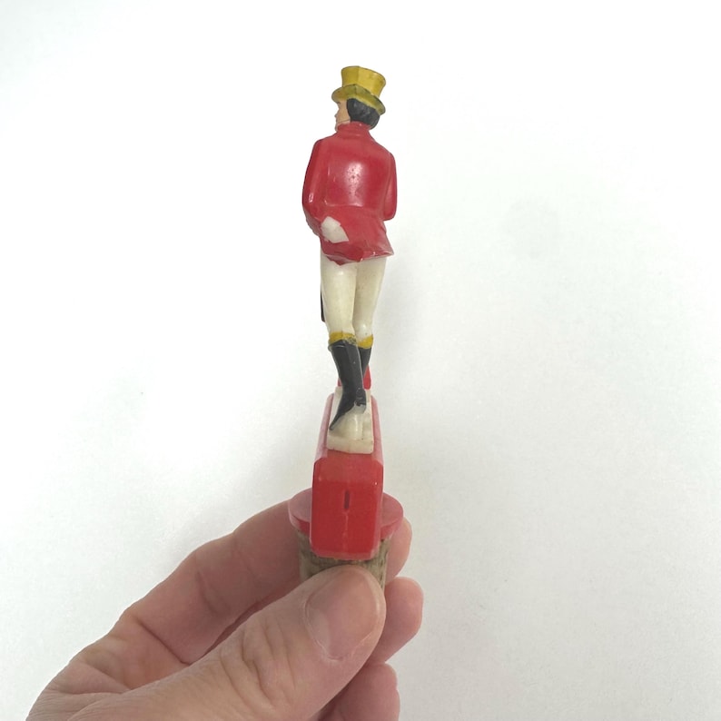 Puede incluir: Un tap&oacute;n de botella vintage con una figura de una persona con abrigo rojo, sombrero de copa amarillo y botas negras. La figura est&aacute; de pie sobre una base rectangular roja. El tap&oacute;n se sostiene en una mano.