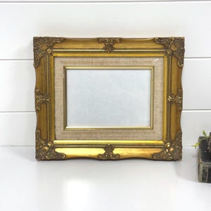 May include: A rectangular picture frame with an ornate, metallic gold finish featuring decorative corner and edge detailing. The frame includes a light beige, textured fabric matting surrounding a central rectangular opening for a photograph.