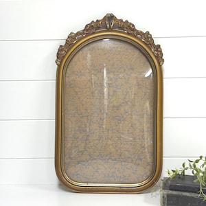 May include: An arched gold picture frame with an ornate, decorative top featuring carved leaf details. The frame contains a glass pane covering a tan backing material that has a faint, repeating blue marbled or vine-like pattern printed on it.
