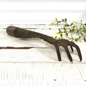 May include: An antique garden rake with a wooden handle and a metal head. The handle is a light brown color, and the metal head has a weathered, green-brown patina. The rake has four tines and is designed for gardening.