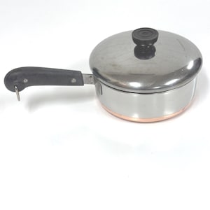 May include: A stainless steel saucepan with a copper-colored base and a matching lid. The pan features a black heat-resistant handle with a small metal hanging loop at the end and a black knob on the center of the lid for easy lifting.