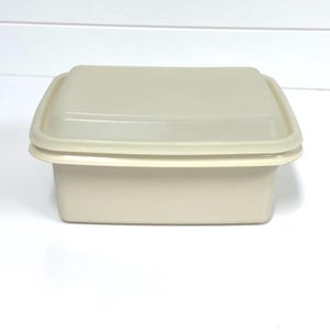 May include: A rectangular, cream-colored plastic food storage container with a matching lid. The container features a smooth, solid-colored finish with a slightly raised rim around the top edge where the lid secures to the base, creating a simple, functional design for kitchen organization.