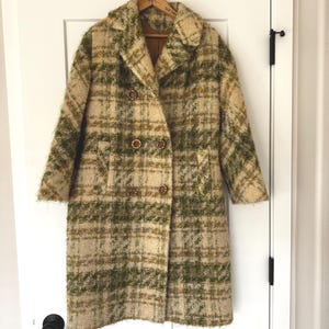 May include: A vintage double-breasted coat in a plaid pattern of green, tan, and cream. The coat has a wide collar, two rows of brown buttons, and long sleeves. It hangs on a wooden hanger against a white door.
