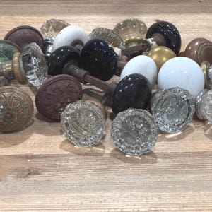 May include: A collection of antique door knobs in various styles and materials. The knobs include clear glass, white, black, and brown knobs with ornate and simple designs. Some knobs have brass or bronze hardware.