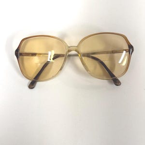 May include: Vintage oversized eyeglasses with a light brown frame and tinted lenses. The glasses have a classic, retro design with a rectangular shape and dark brown temples. The lenses are a warm, amber color.