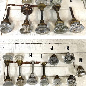 May include: A collection of vintage glass door knobs with brass and metal hardware. The knobs have various shapes and designs, including faceted and round styles. The background is a distressed white wooden surface.