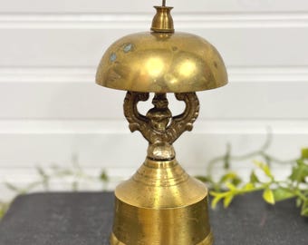 May include: A vintage brass bell with a decorative handle and a round top. The bell is on a dark brown surface.