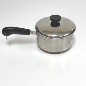 May include: Stainless steel saucepan with a black handle and lid. The lid has a black knob. The saucepan is on a white surface. This kitchenware is ideal for cooking.